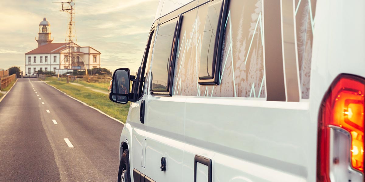 A white converted campervan driving on the road towards a lighthouse