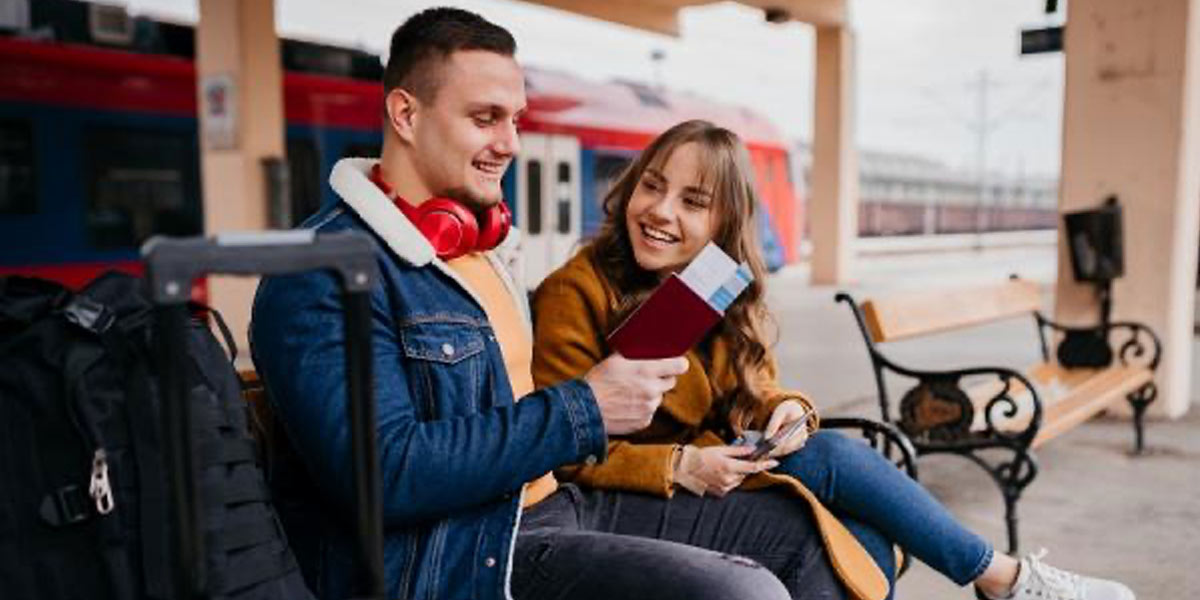 A smiling couple sat on a bench at the train station looking at their travel documents
