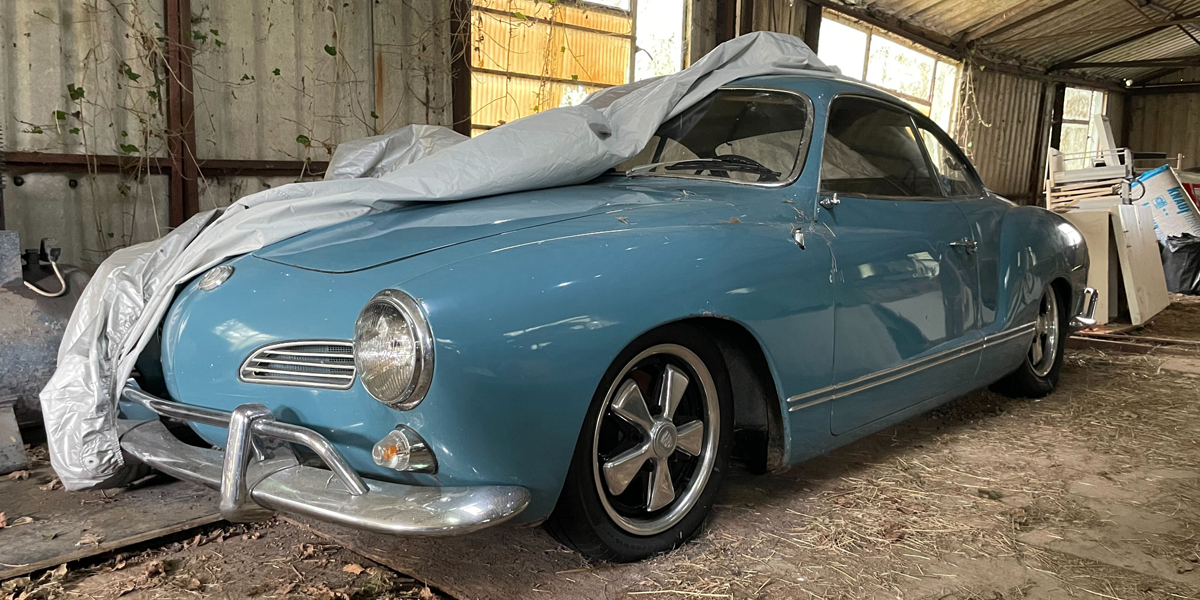A VW Karmann Ghia parked up in a storage barn, with its vehicle cover rolled back